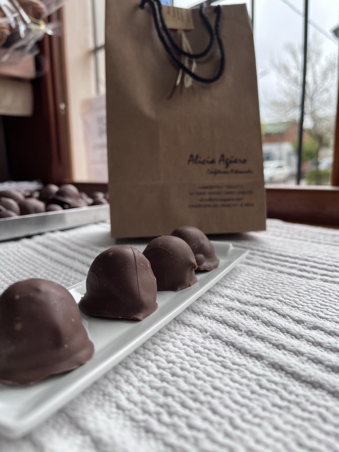 Bombones de chocolate bañados a mano sobre un plato blanco, junto a una bolsa de papel con el logo de Alicia Agüero